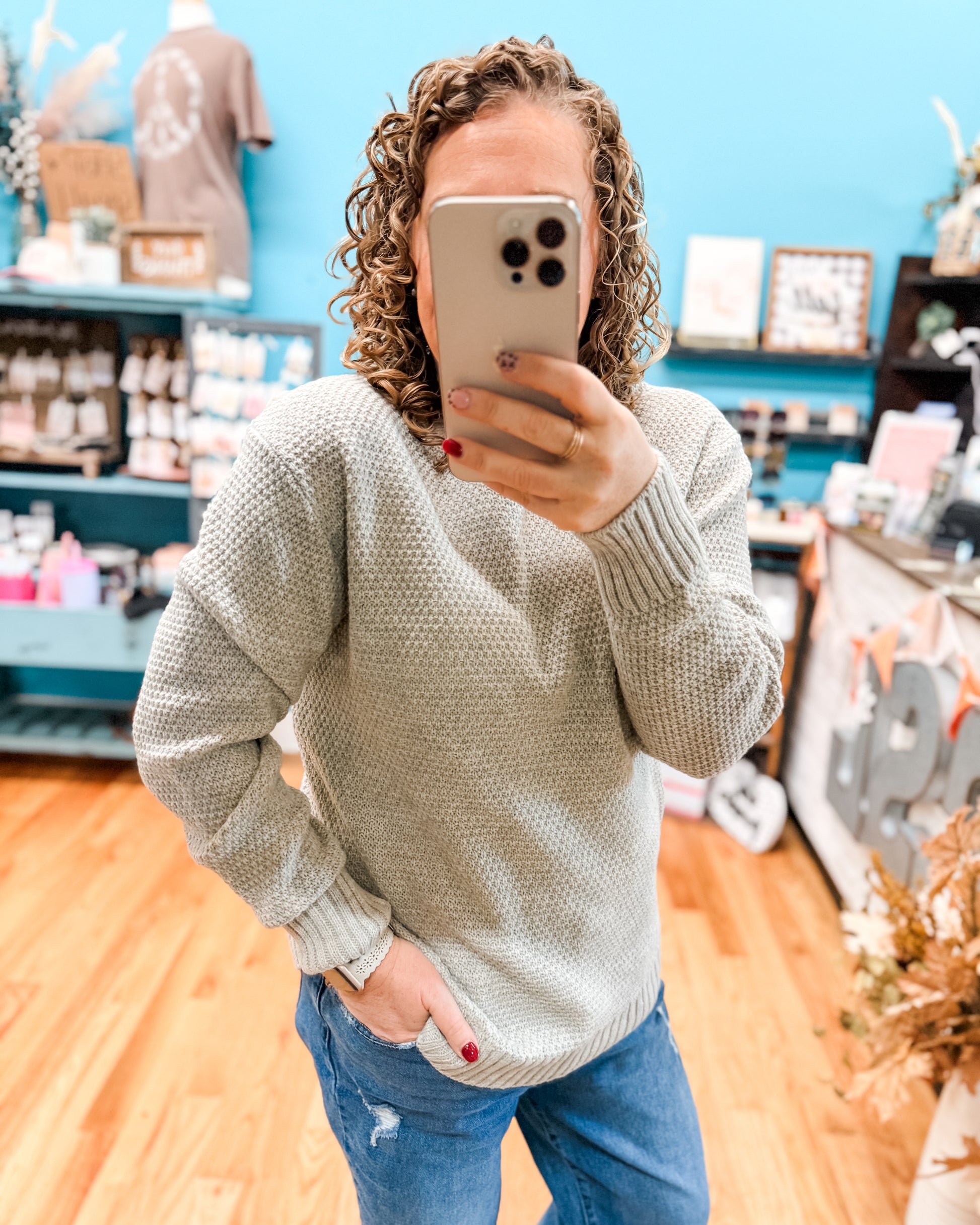 Person wearing a light gray sweater and blue jeans taking a mirror selfie in a store.