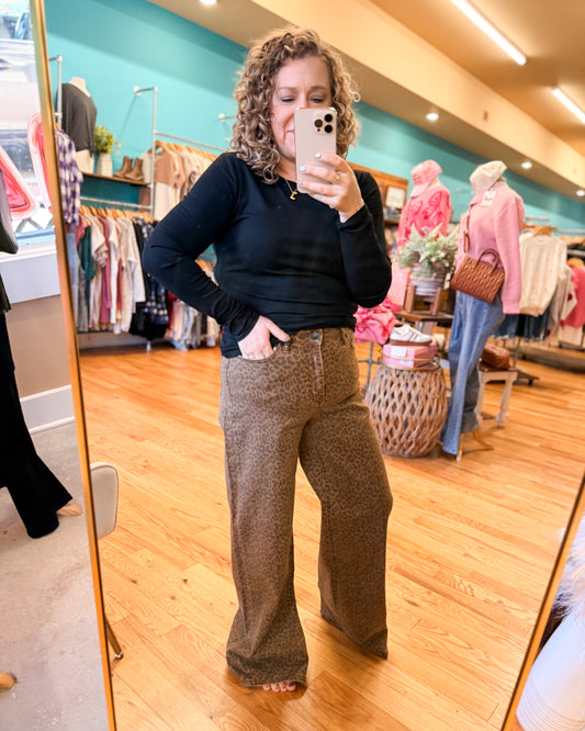 Woman taking a mirror selfie in a clothing store wearing a black top and brown patterned pants.