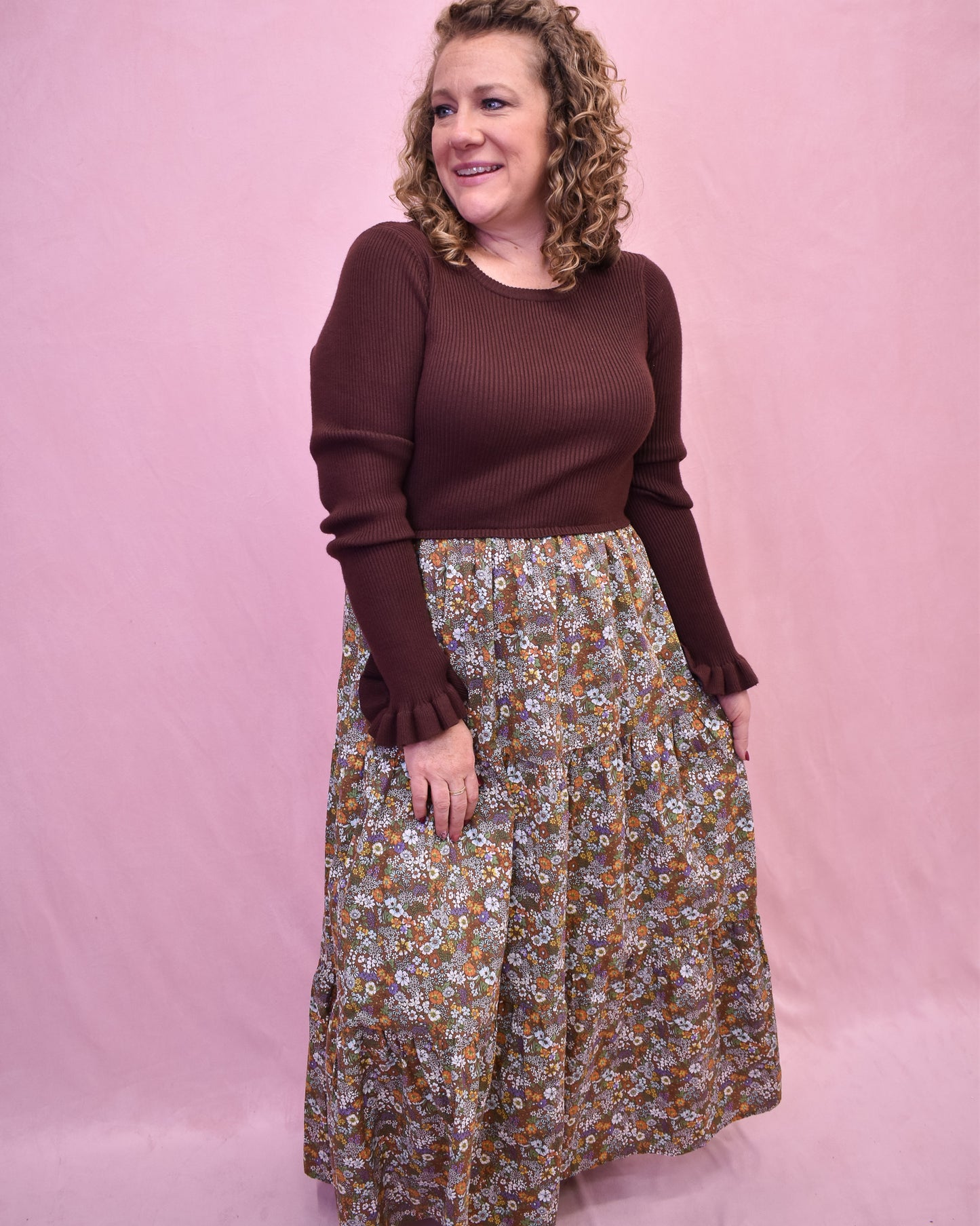 Woman wearing a brown sweater and floral skirt against a pink background