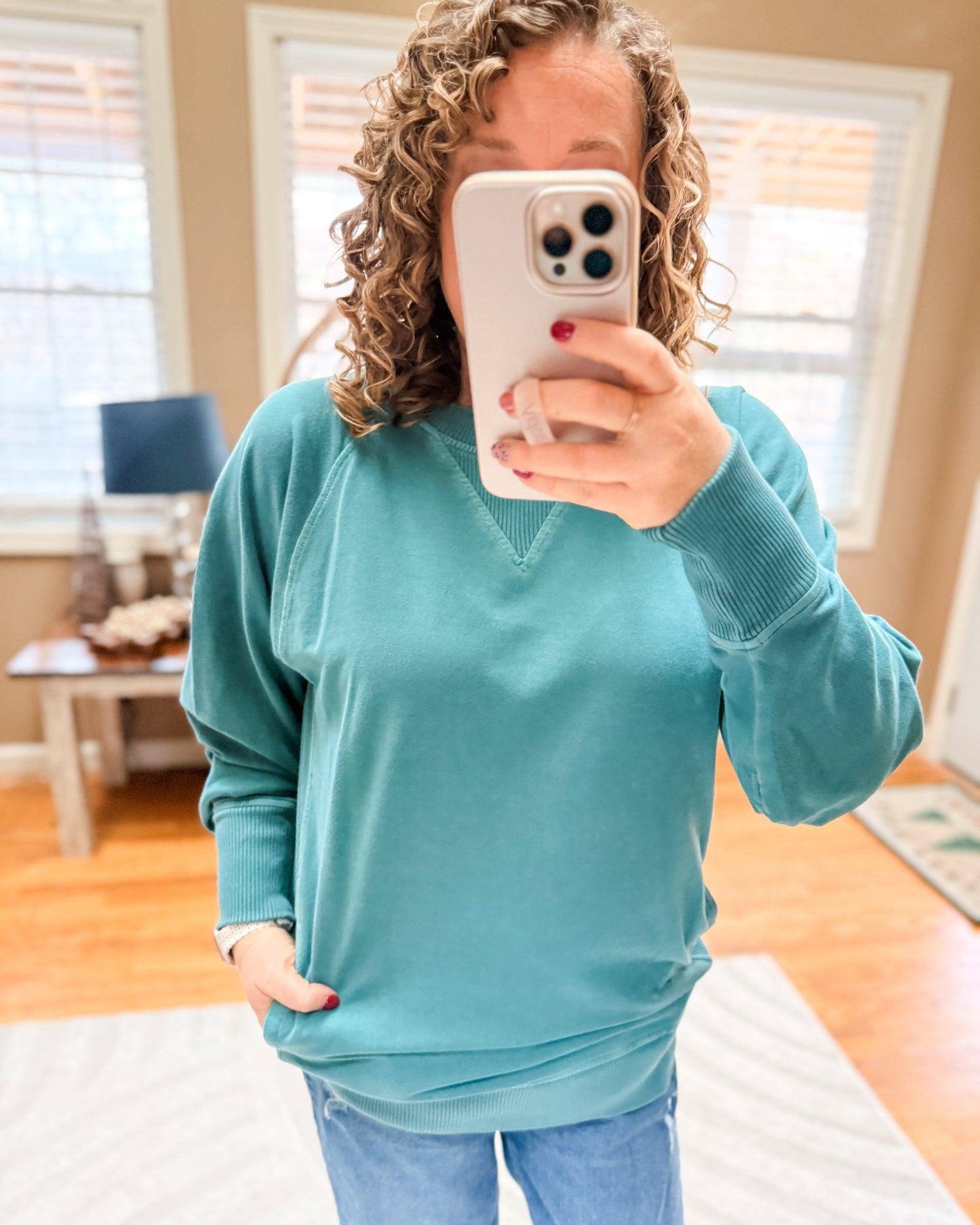 Person wearing a teal sweater taking a mirror selfie in a room with wooden floors and a window.