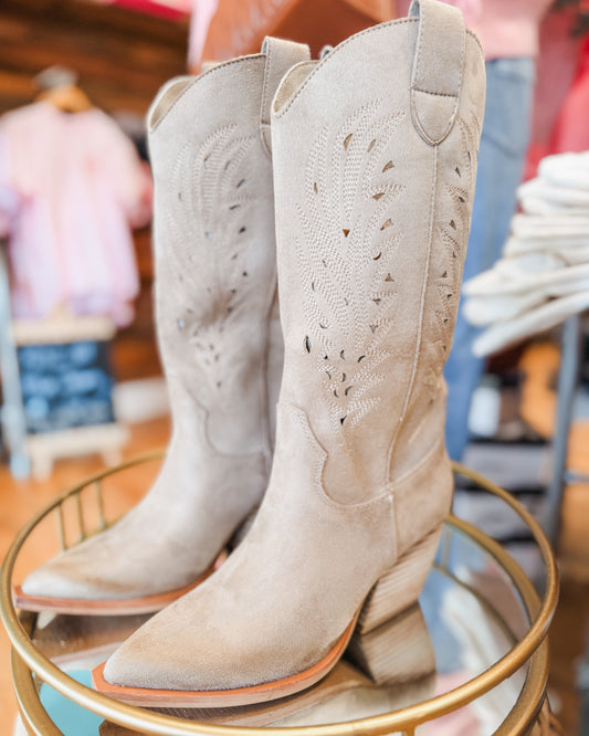 Pair of beige cowboy boots on a round metal stand with a blurred indoor background.
