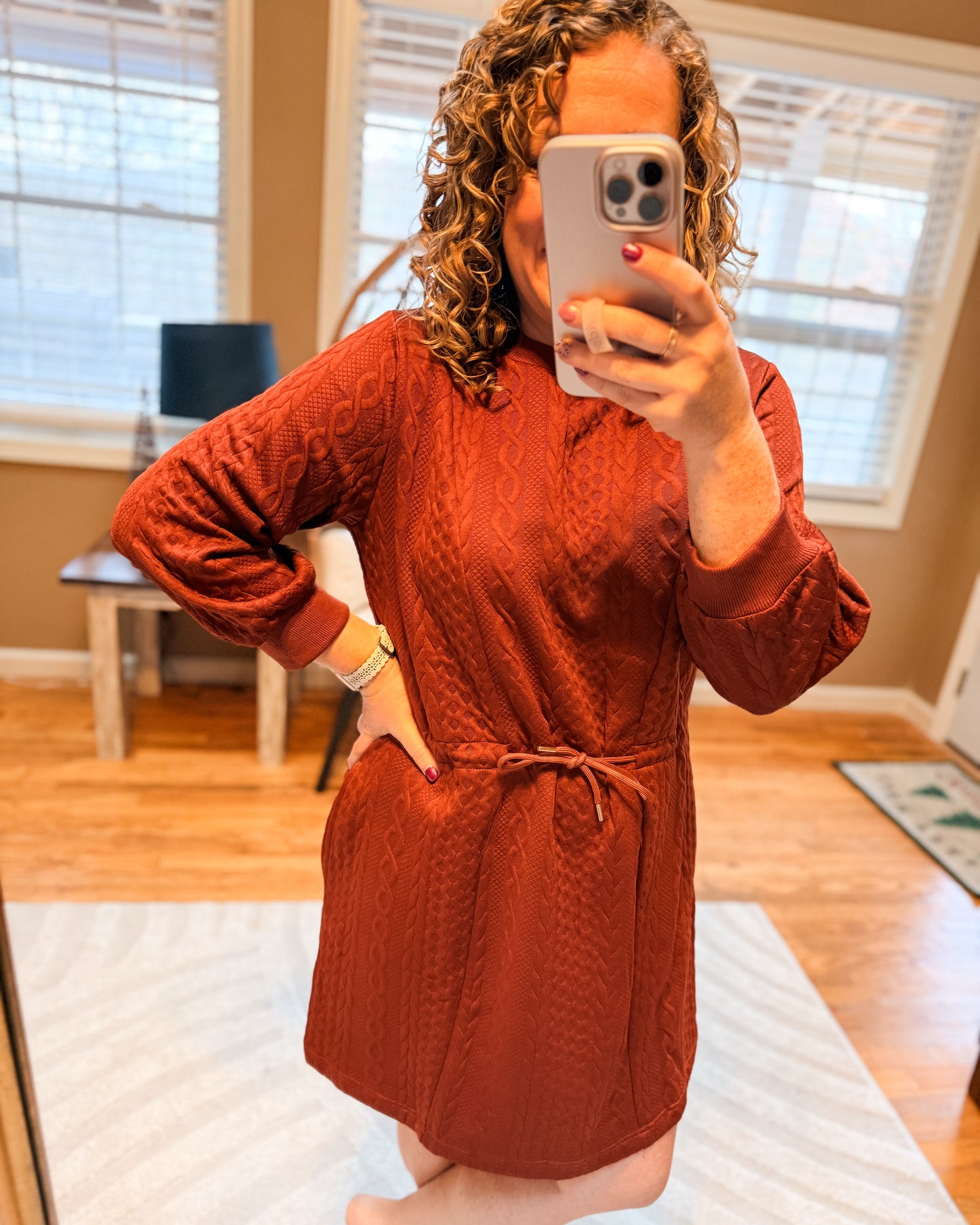 Person wearing a rust-colored robe taking a mirror selfie in a room with wooden flooring and windows.