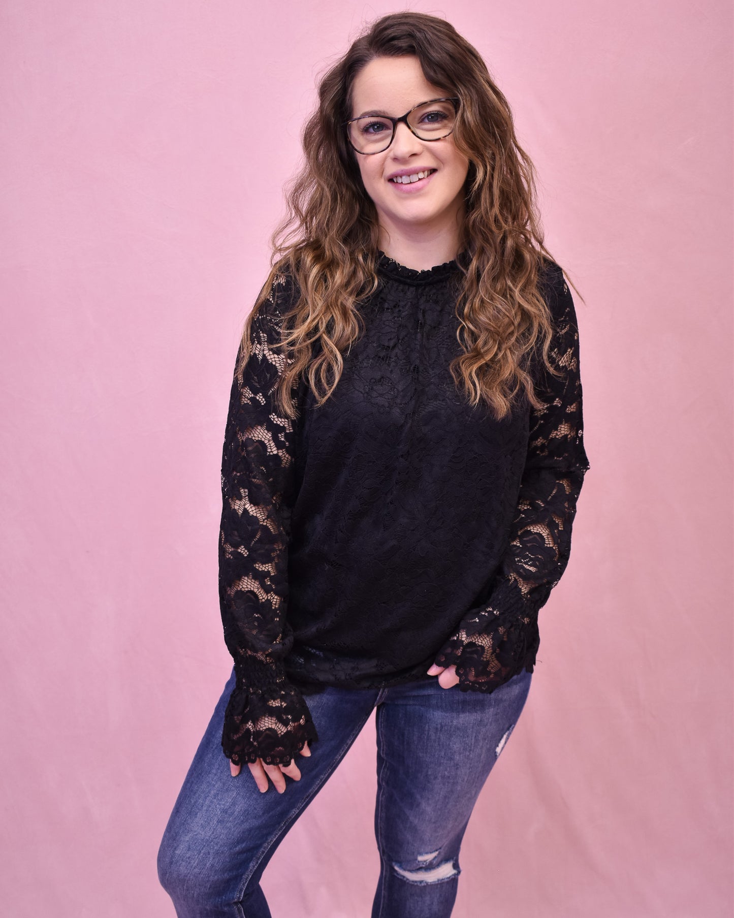 Woman wearing a black lace-sleeve top and jeans against a pink background
