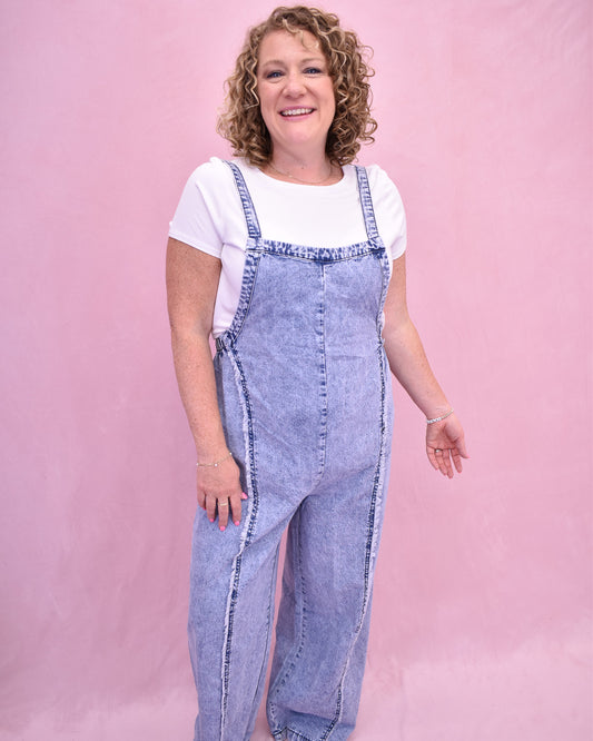 Woman wearing denim overalls and a white shirt against a pink background
