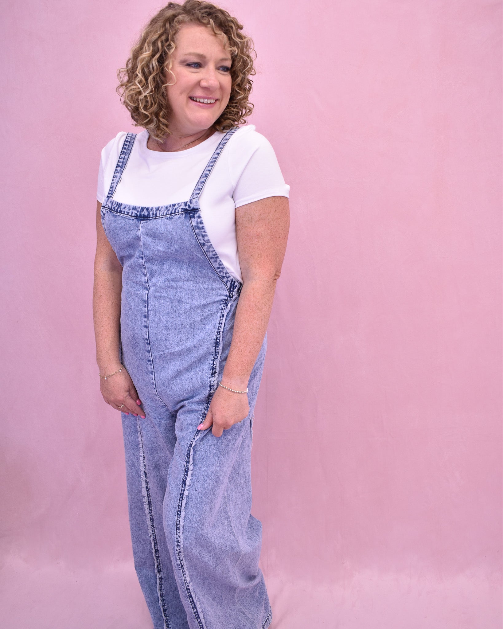 Person wearing a denim jumpsuit over a white shirt on a pink background