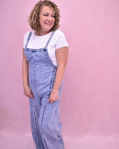 Person wearing a denim jumpsuit over a white shirt on a pink background