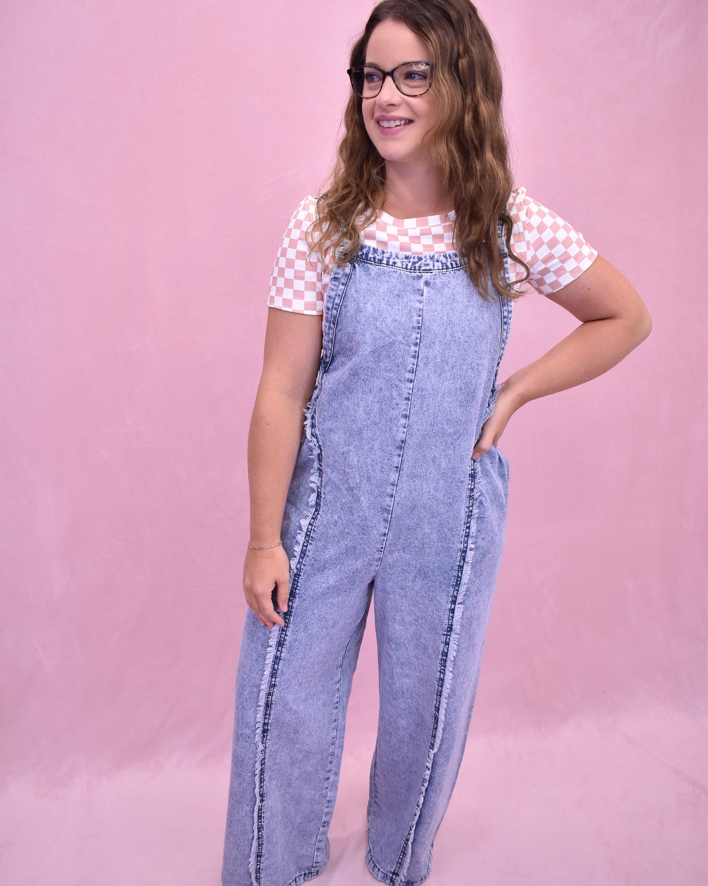 Woman wearing denim overalls and a checkered shirt against a pink background