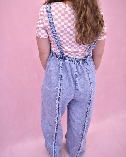 Person wearing denim overalls and a checkered shirt on a pink background