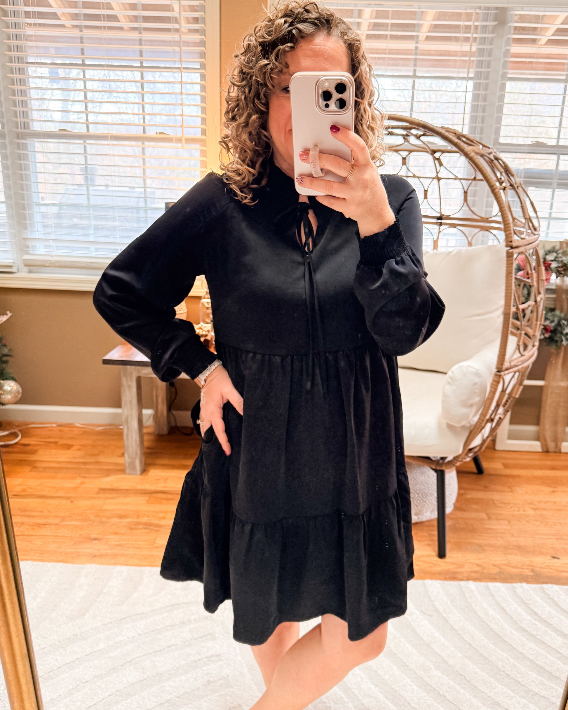 Person wearing a black dress taking a mirror selfie in a room with wooden flooring and a wicker chair.