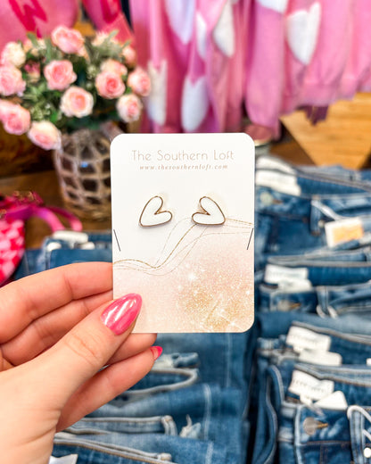 Heart-shaped earrings on a card held by a hand with pink nail polish, with denim jeans and flowers in the background.