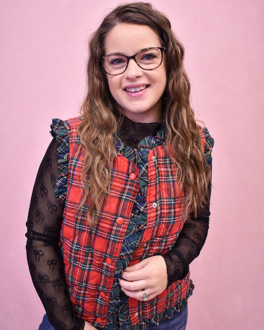 Person wearing a red plaid vest over a black lace top against a pink background