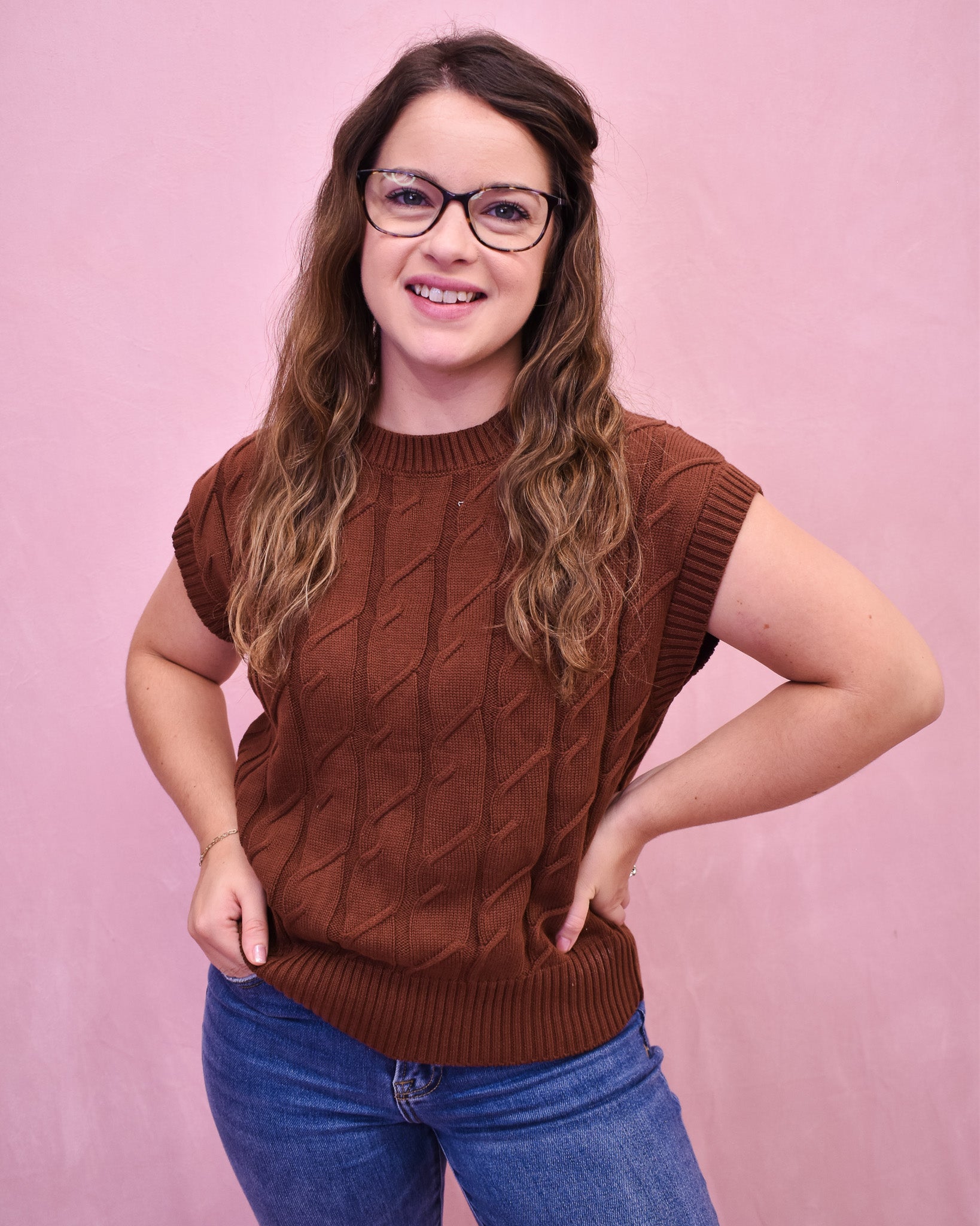 Woman wearing a brown cable knit sleeveless sweater against a pink background