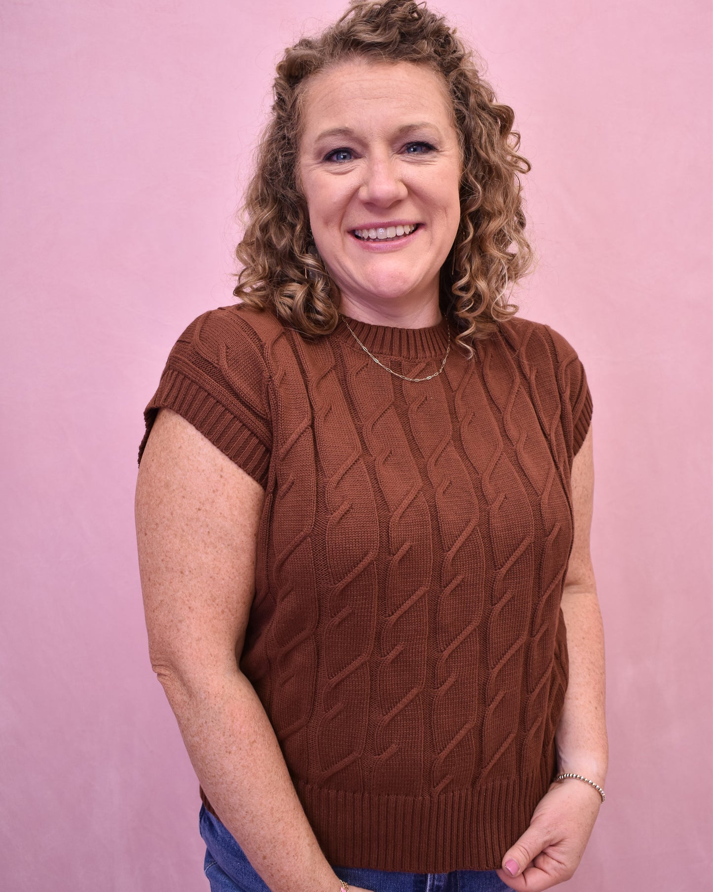Woman wearing a brown sleeveless sweater against a pink background