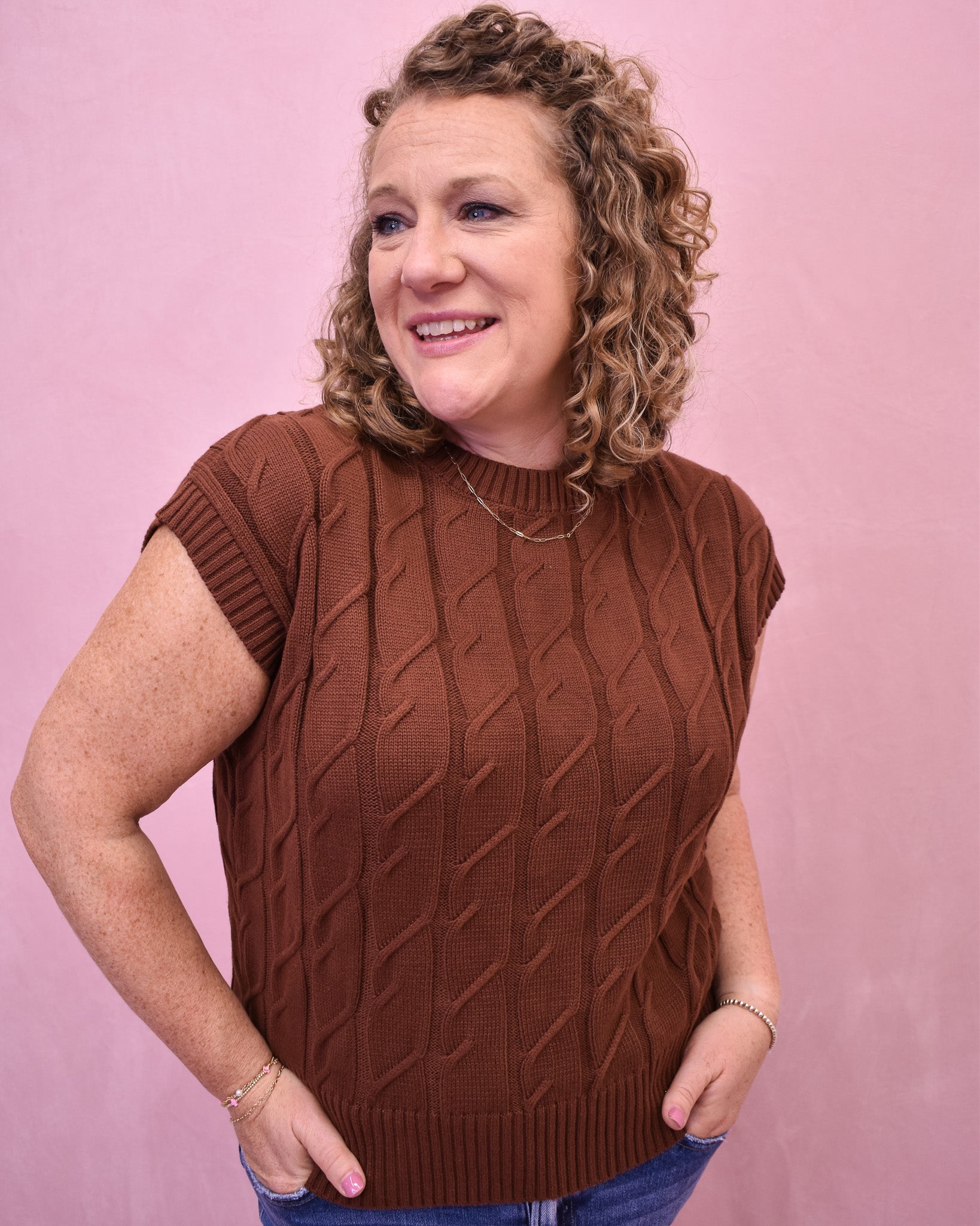 Woman wearing a brown cable knit sleeveless sweater against a pink background
