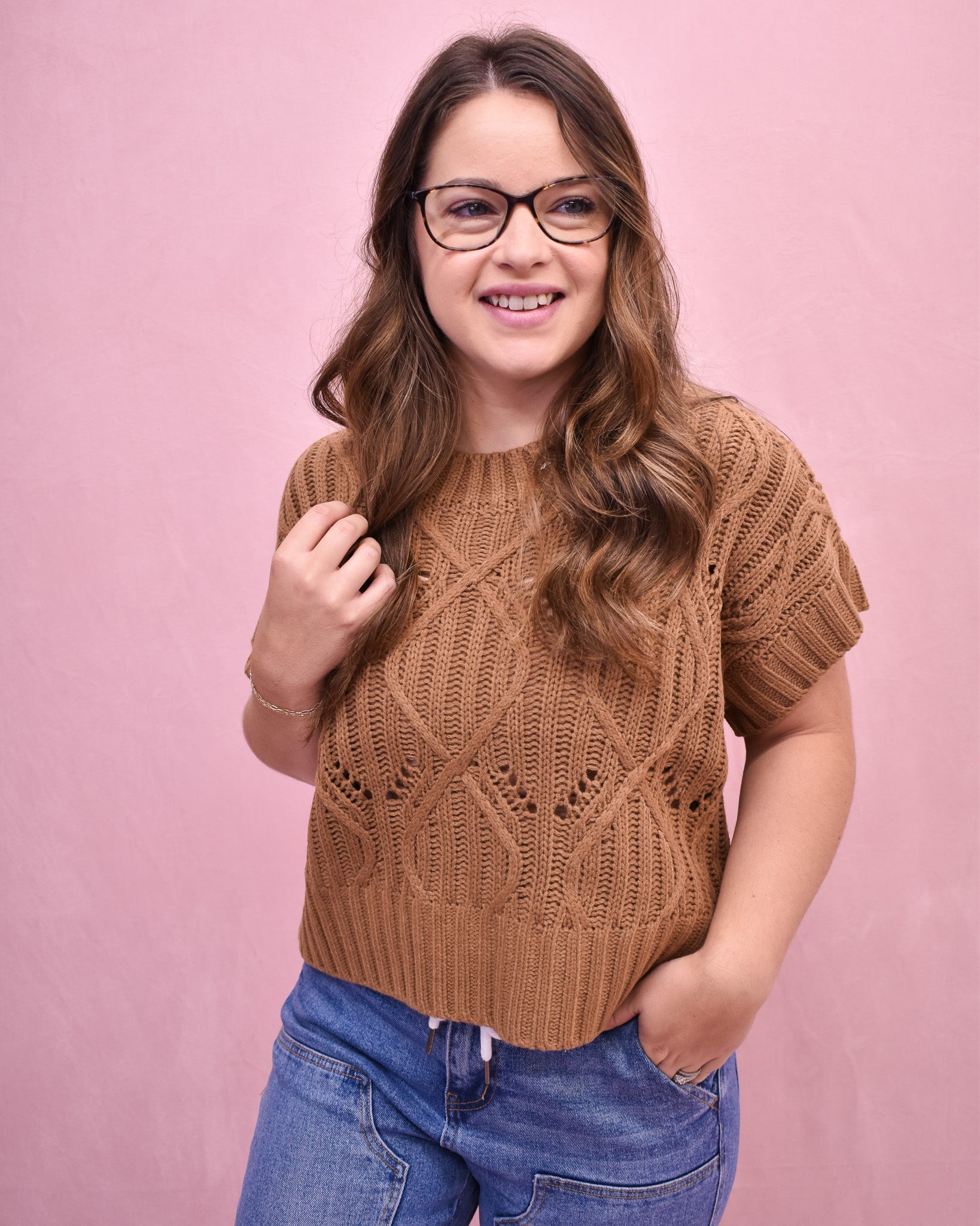 Woman wearing a brown knitted sweater and blue jeans against a pink background