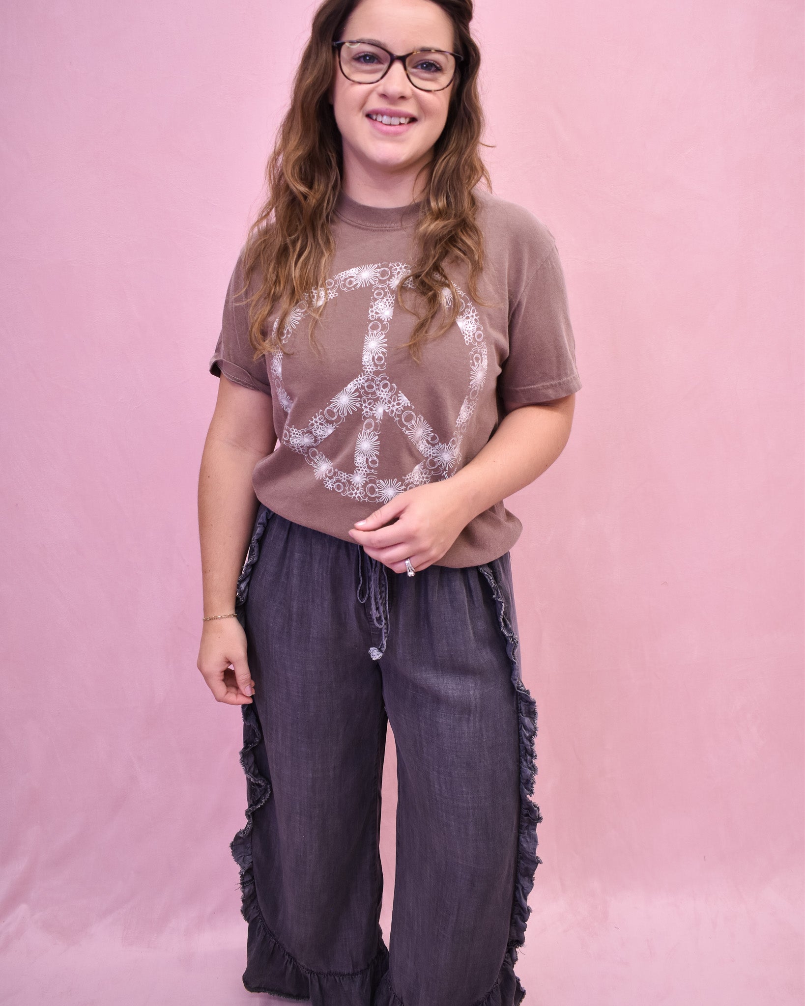 Person wearing a brown t-shirt with a peace symbol design and dark pants against a pink background